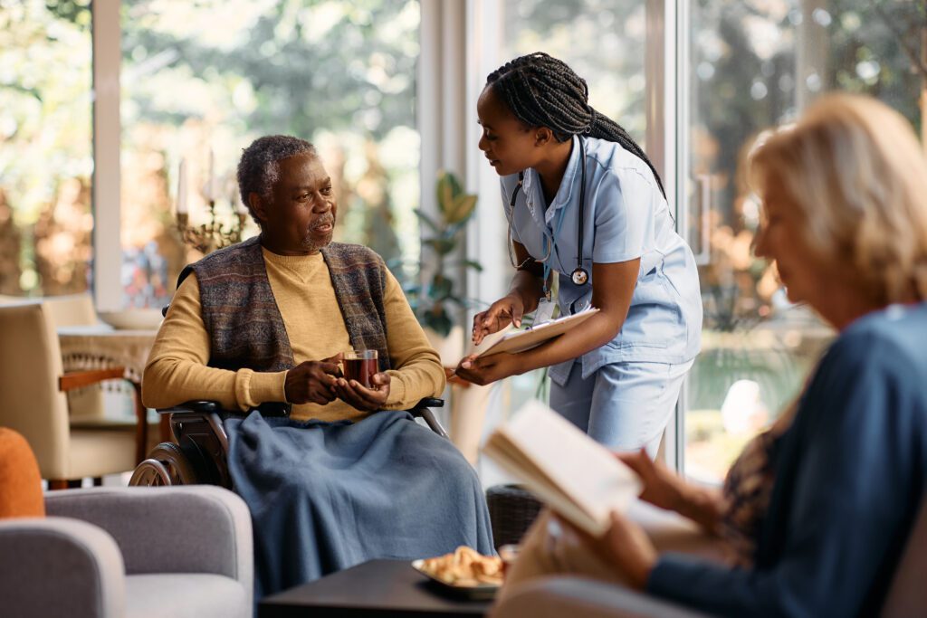 Black senior man in wheelchair drinking tea while talking to a nurse at residential care home.